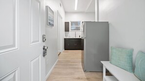 Un pasillo estrecho con una puerta blanca, un banco con cojines color turquesa y una cocina al fondo.