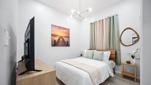 Un dormitorio moderno con una cama, un televisor sobre un escritorio de madera, un cuadro, cortinas, un espejo y una planta pequeña.