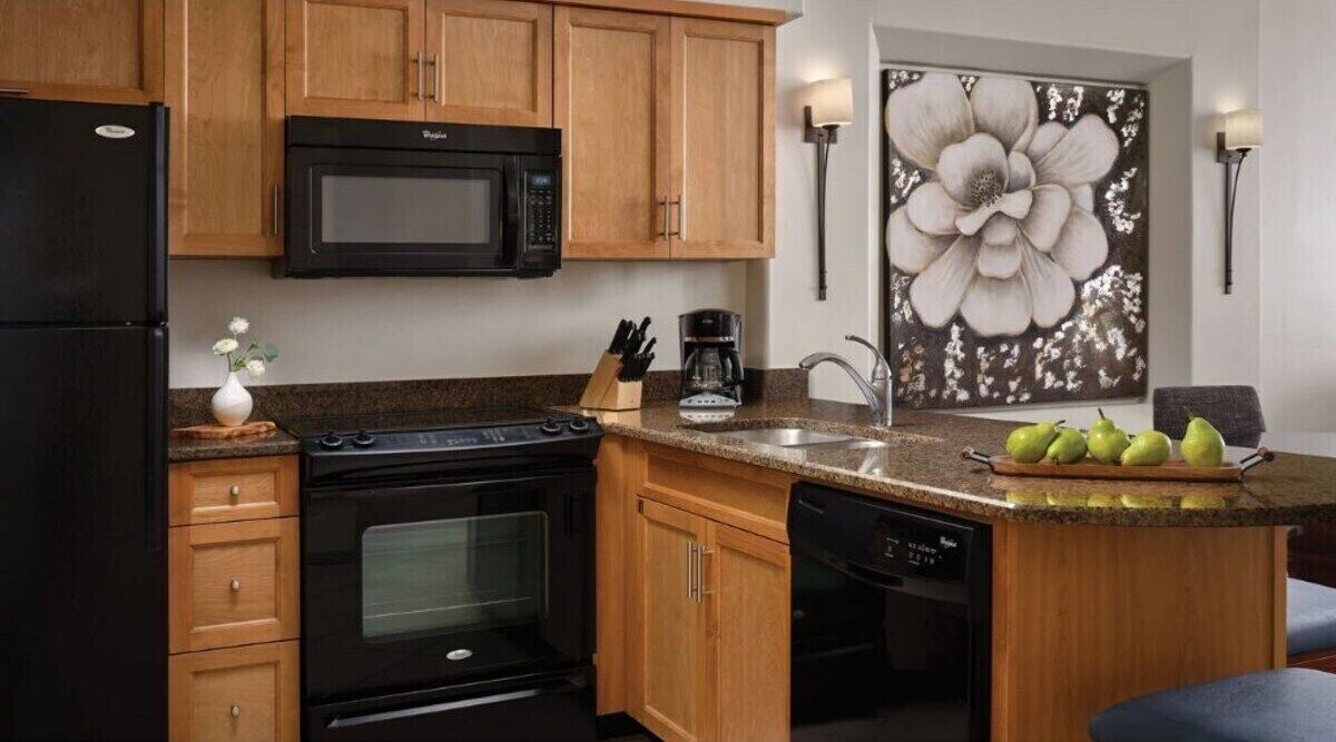 A kitchen with wooden cabinets, a black refrigerator, a microwave, an oven, a dishwasher, a granite countertop, a coffee maker, a sink, and a large floral artwork on the wall.