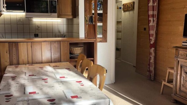 A dining area with a table covered in a tablecloth with heart patterns, wooden chairs, a wooden cabinet, and a microwave above the counter. - Charming 2-room Apartment for 4/6 People (Bessans)
