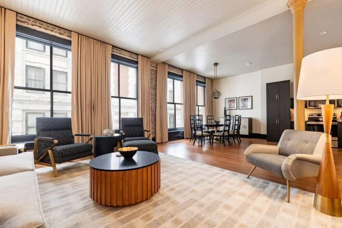 A spacious living room with a round coffee table, two armchairs, a sofa, a dining area with chairs, and large windows with curtains.