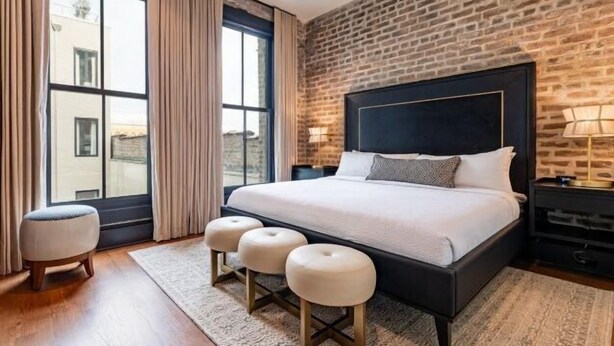 A bedroom with a brick wall, a large bed with a headboard, a window with curtains, and a nightstand with a lamp. - 2BR Elegant French Quarter Escape The Mayfair (New Orleans)