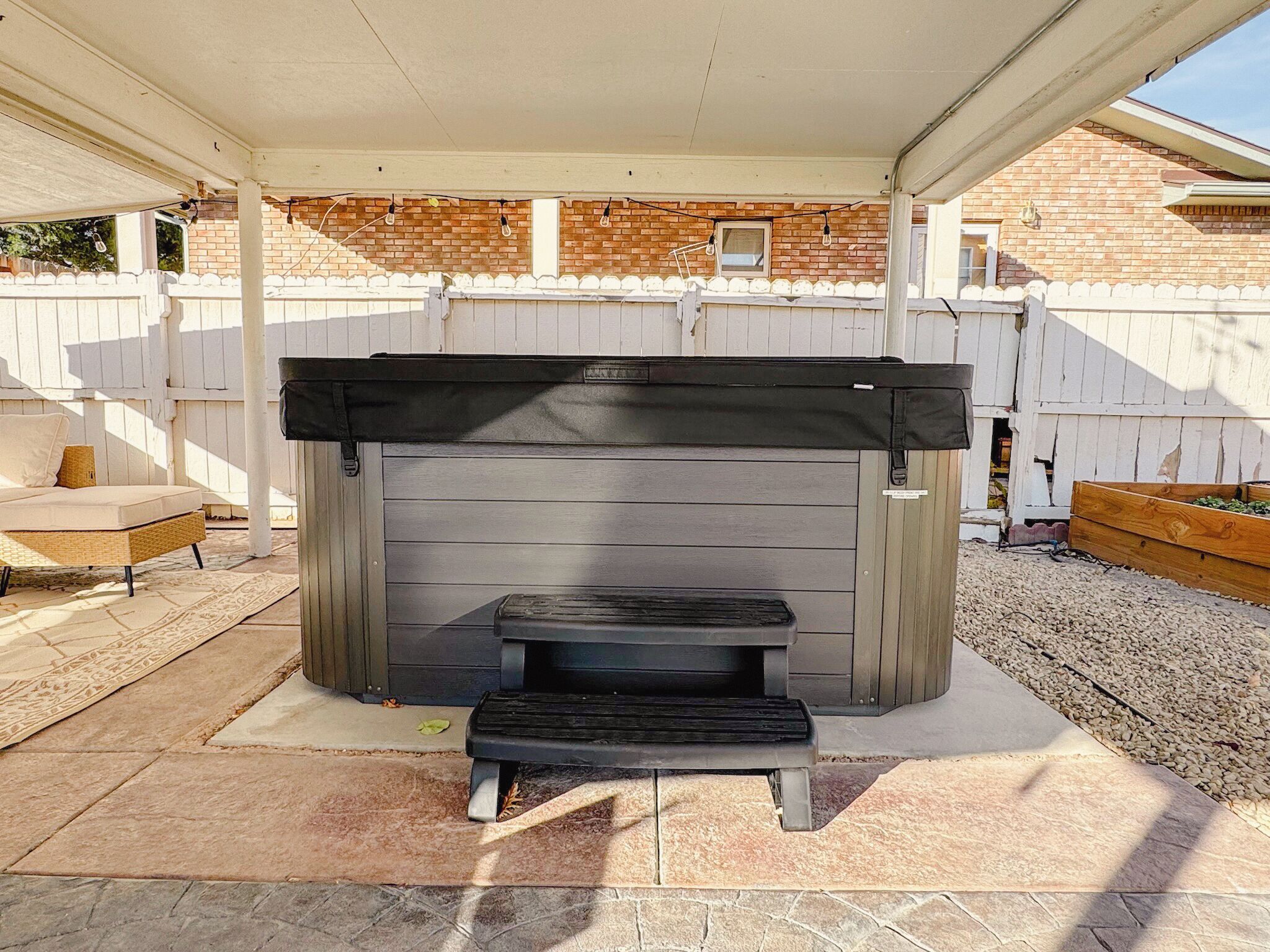 A covered outdoor grill area with a bench and a small table, surrounded by a white fence and a brick wall.