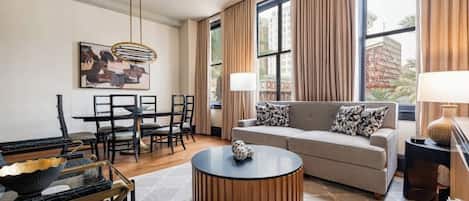 A modern living room with a round coffee table, a sofa with patterned cushions, a dining area with chairs, and a large window with curtains.