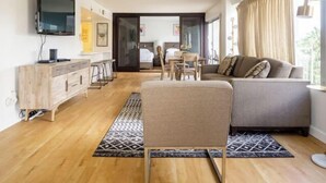 A modern living room with a sofa, a TV, a wooden cabinet, and a dining area with chairs. - City & Palm View Oceanfront Bliss w/ Free Parking (Honolulu)