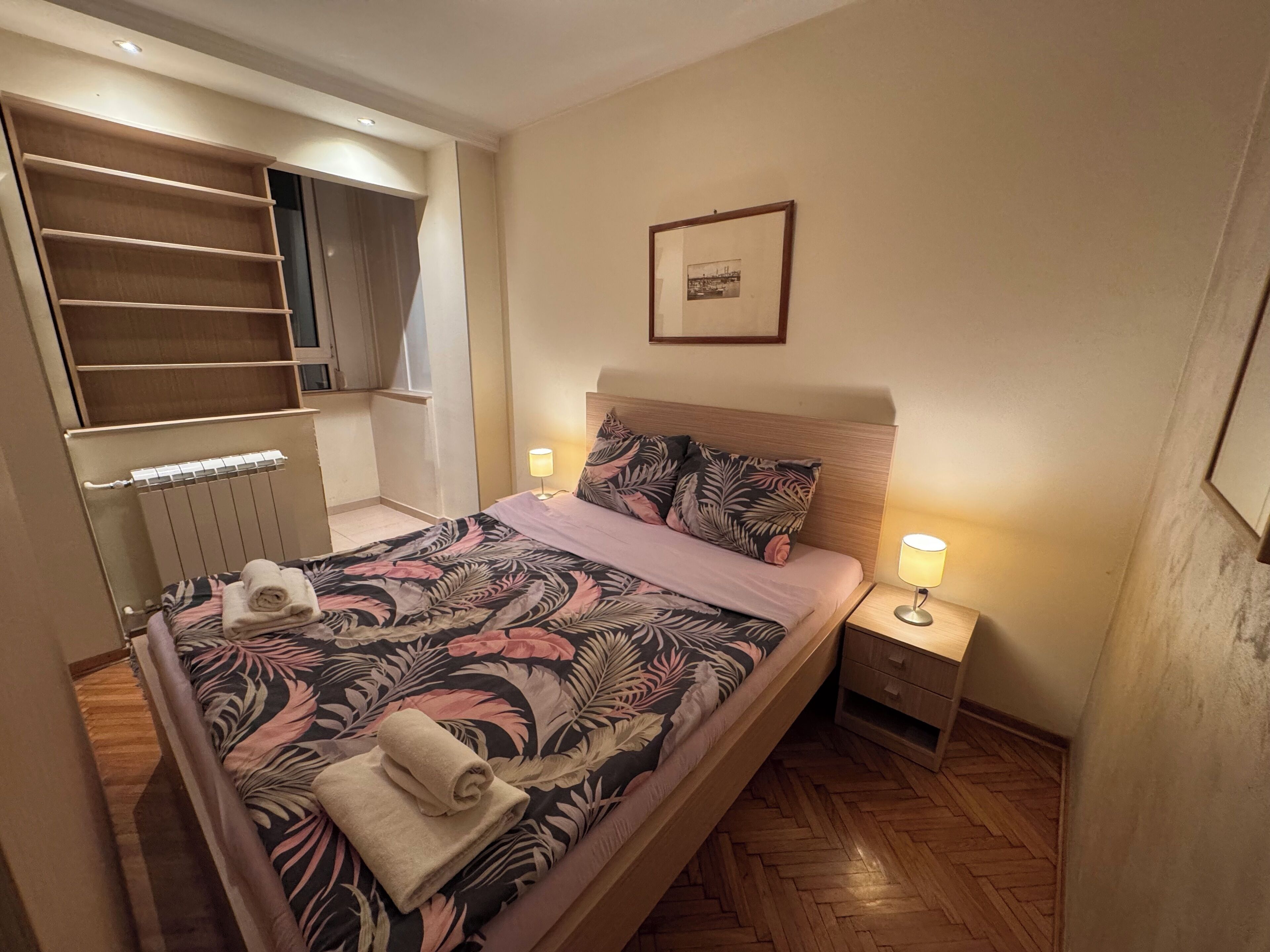A bedroom with a bed, two bedside lamps, a radiator, a wardrobe, and a framed picture on the wall.