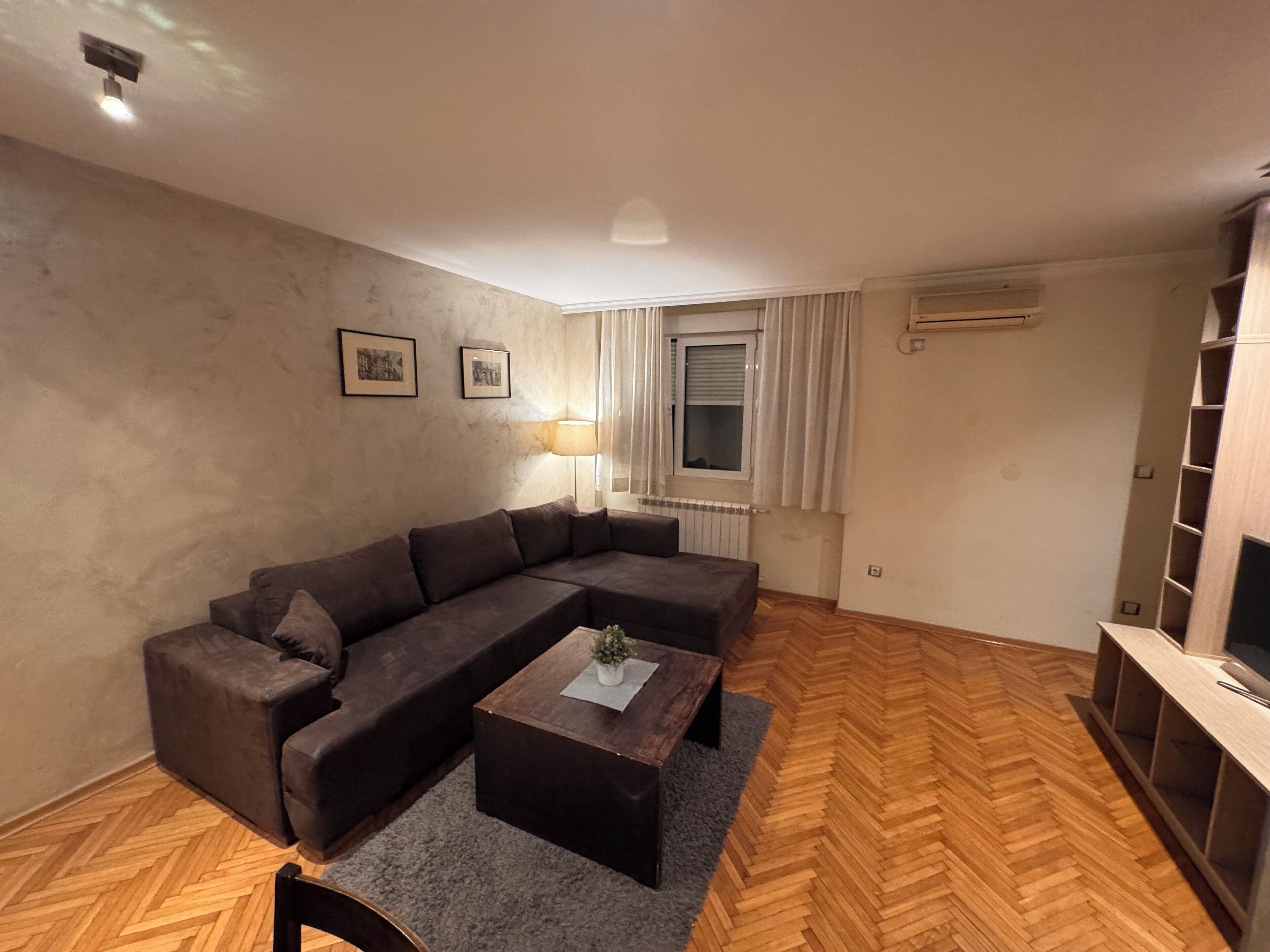 A living room with a sectional sofa, a wooden herringbone floor, a small coffee table, a lamp, and a TV stand.