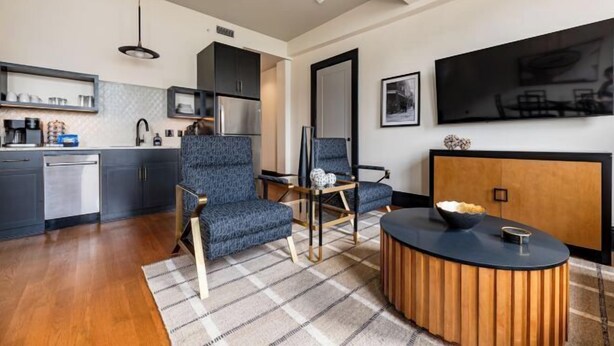 Una sala moderna con un sofá, sillas, una mesa de centro y una cocina con armarios y electrodomésticos.