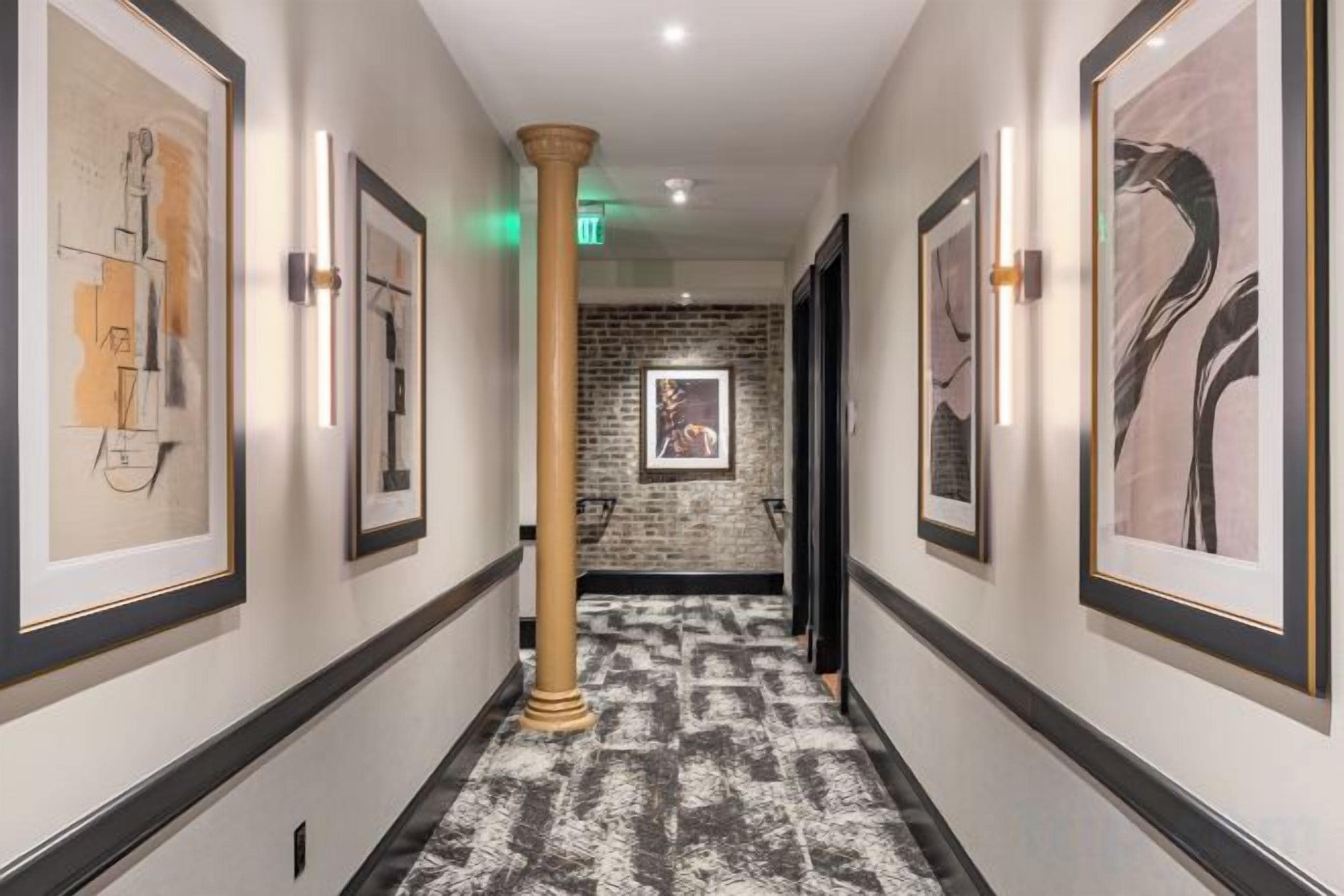 A hallway with framed artwork on the walls, a patterned carpet, and a brick wall in the background.