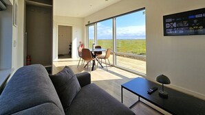 A modern living room with a sofa, a dining table with chairs, and a large window offering a view of the outdoors. - Hítarneskot – Private Hot Tub Cottage in Snæfellsnes (Borgarnes)