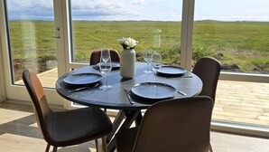 A dining table set for four with a view of a grassy field through large windows. - Hítarneskot – Private Hot Tub Cottage in Snæfellsnes (Borgarnes)
