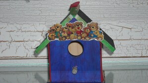 Una casita de pájaros de madera con base azul y techo verde, decorada con pegatinas de ositos de peluche.