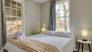 A neatly made bed with a beige blanket, a window with a view of a wooden deck, and a small bedside table with a lamp. - Pet-Friendly Pensacola Home Close to Beaches & Fun (Pensacola)