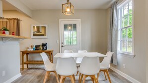 A dining area with a white table and chairs, a wooden side table with kitchen items, a hanging light fixture, and a window with curtains. - Pet-Friendly Pensacola Home Close to Beaches & Fun (Pensacola)