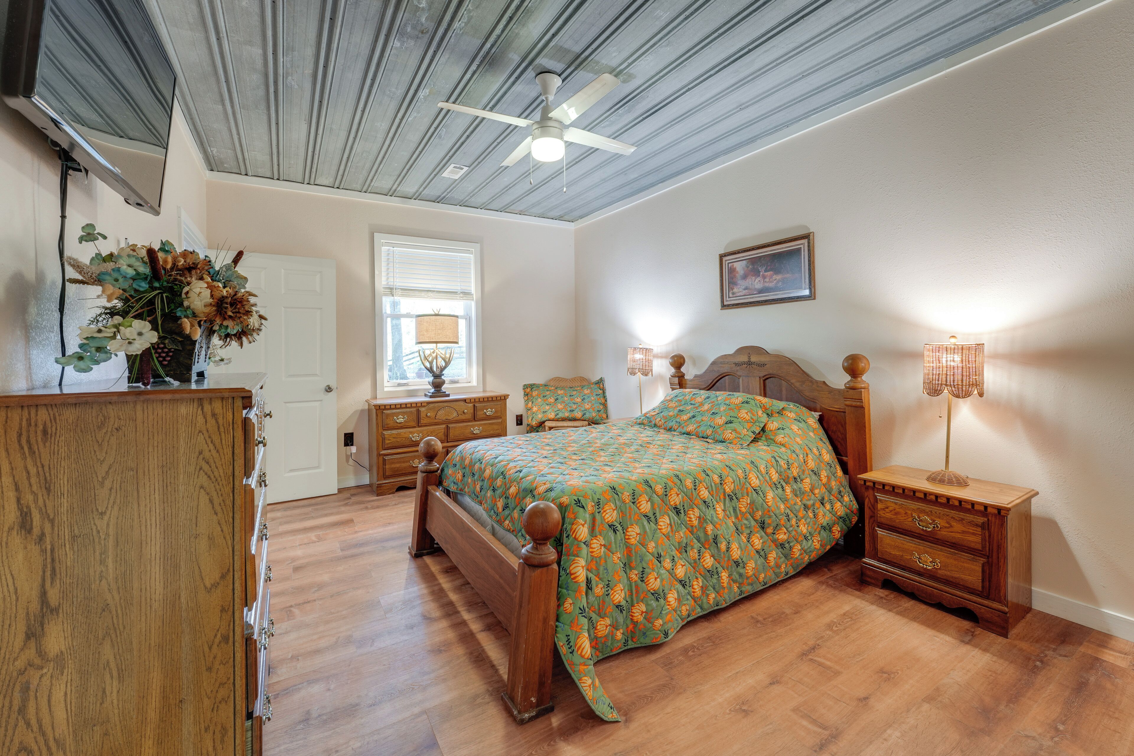 A bedroom with a wooden bed, a nightstand with a lamp, a dresser, a ceiling fan, and a window with blinds.
