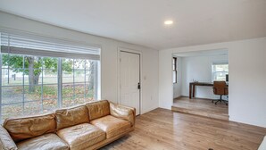Ein geräumiges Wohnzimmer mit Ledersofa, Holzböden, großen Fenstern und einer Tür zu einem anderen Raum.