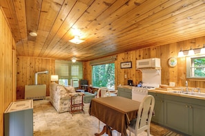 A wooden cabin interior with a dining area, kitchen, and a large window providing a view of the outdoors.