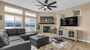A living room with a sectional sofa, a fireplace, a mounted TV, and large windows offering a view of the outdoors. - Lakefront Home w/ Mountain Views • Walk to Shops (Sparks)