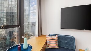 Ein modernes Wohnzimmer mit großem Fenster, einem blauen Sofa, einem farbenfrohen Streifen-Kissen, einem kleinen Tisch und einem Wand-Fernseher.
