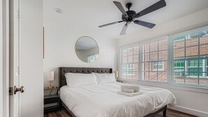 Un dormitorio con cama, ventilador de techo, ventana con persianas, mesita de noche y espejo.