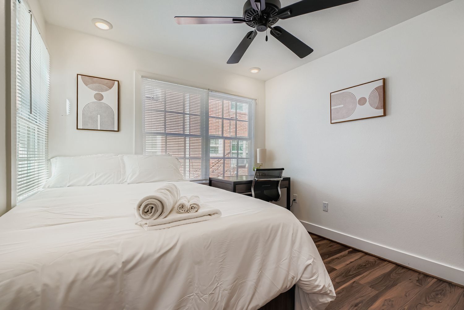 A hotel room with a bed, a desk, a chair, a ceiling fan, and two windows with blinds.