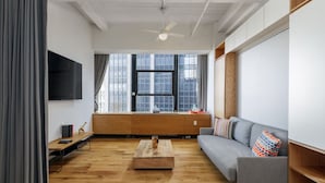 A modern living room with a wooden floor, a grey sofa, a coffee table, and a TV mounted on the wall. - Central FiDi Studio Near NYC’s Top Attractions, Dining, and Iconic Landmarks (New York)