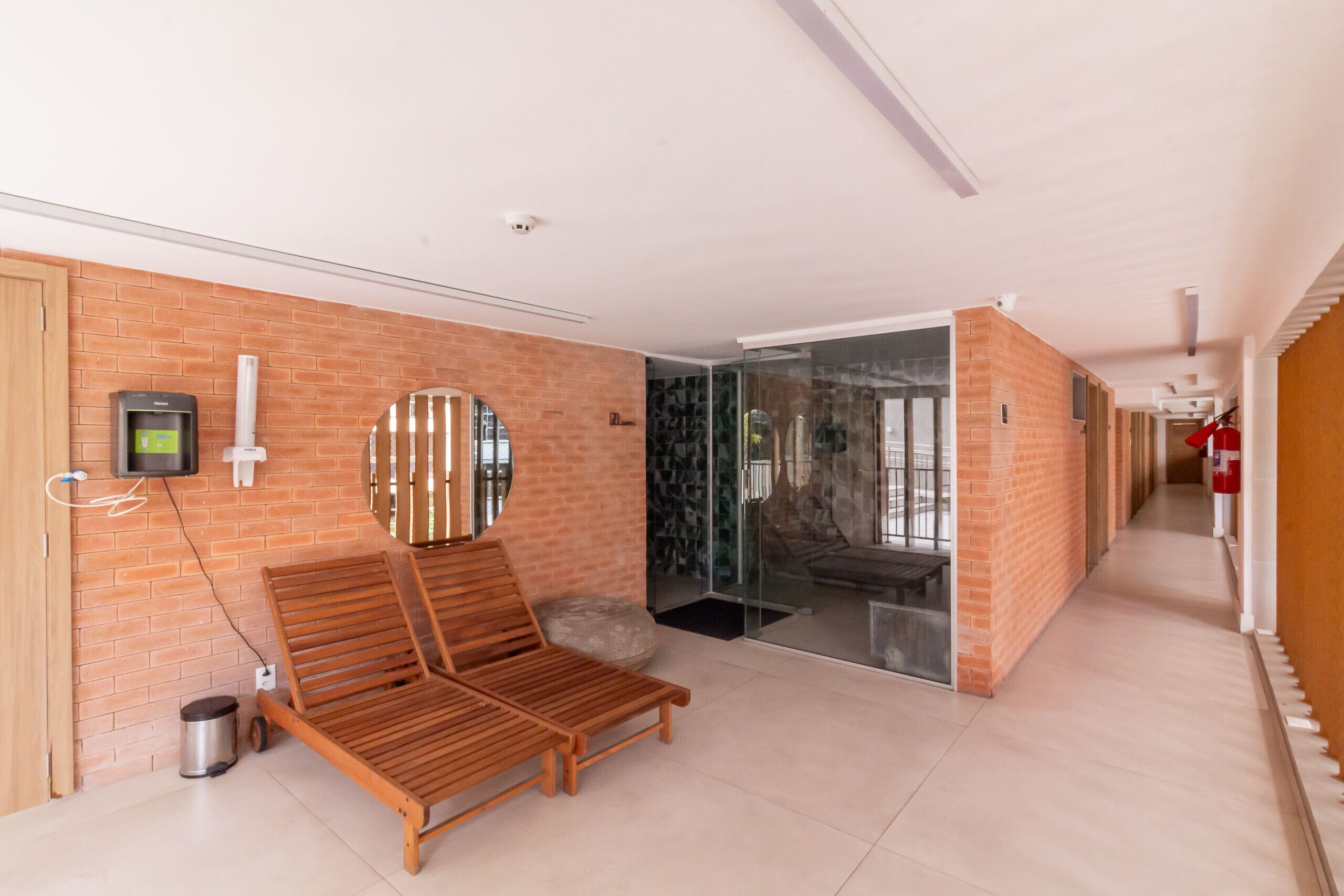 A modern hotel lobby with a wooden lounge area, a glass door leading to a room, and a brick wall with a mounted device.