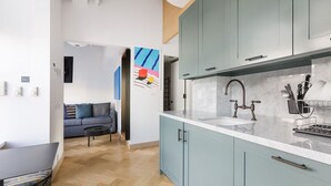 Una cuina moderna amb armaris de blau clar, un taulell blanc i un rentaplat. Hi ha una zona de menjador amb taula i cadires, i un sofà al fons.