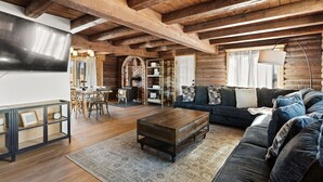 A rustic log cabin interior with a wooden ceiling, a living room with a sofa and coffee table, a dining area with a table and chairs, and a brick fireplace.