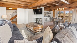 A spacious living area with a wooden ceiling, a large sofa set, a coffee table, and a television mounted on the wall.