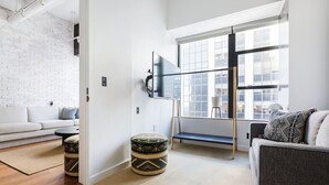 Un salon moderne avec un mur en briques blanches, une grande fenêtre, un téléviseur fixé au mur, un canapé, une table basse et un pouf à motifs.