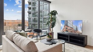Un salon moderne avec une grande fenêtre, un canapé, un fauteuil, une table basse, un téléviseur et une plante en pot.