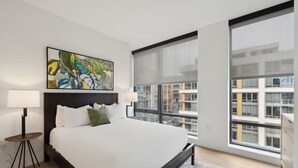 Un dormitorio moderno con una cama grande, una lámpara de pie, un cuadro y vistas a la ciudad a través de amplios ventanales.