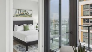 Habitación de hotel moderna con una cama grande, una mesa de comedor con sillas y vistas a la ciudad a través de ventanales de gran altura.