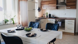 A modern kitchen with a dining table set for two, featuring a white tablecloth, black chairs, and a fruit bowl.
