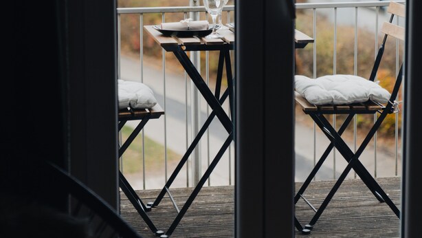 A balcony with a table set for two, a chair, and a window with a view of a building and trees.
