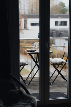 Un balcon avec une table dressée pour deux personnes, une chaise et une fenêtre donnant sur un immeuble et des arbres.