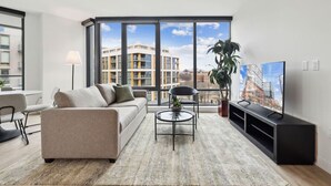Un salon moderne comprenant un canapé, une table basse, un téléviseur et une grande fenêtre donnant sur des immeubles.