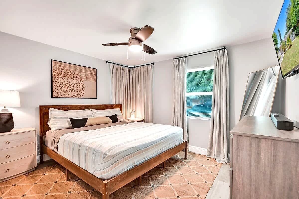 Un dormitorio con cama, ventilador de techo, ventana con cortinas, mesita de noche y televisor sobre un soporte.