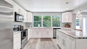Une cuisine moderne avec des meubles blancs, des appareils en acier inoxydable et une grande fenêtre donnant sur un paysage verdoyant.