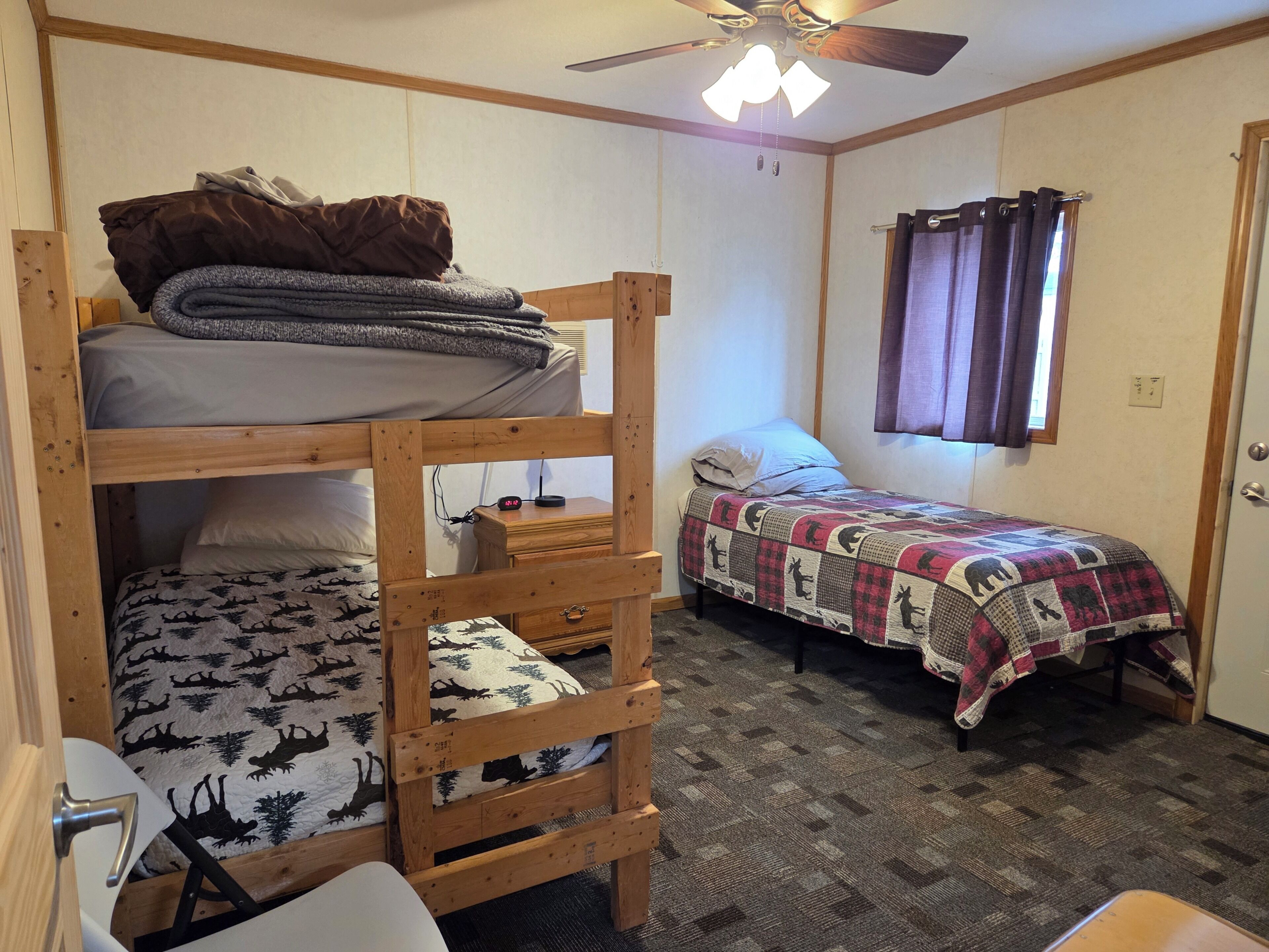 A bunk bed with a desk, a single bed with a patterned blanket, a quilted bed, a ceiling fan, and a window with curtains.