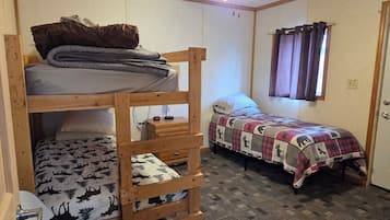 A bunk bed with a desk, a single bed with a patterned blanket, a quilted bed, a ceiling fan, and a window with curtains.