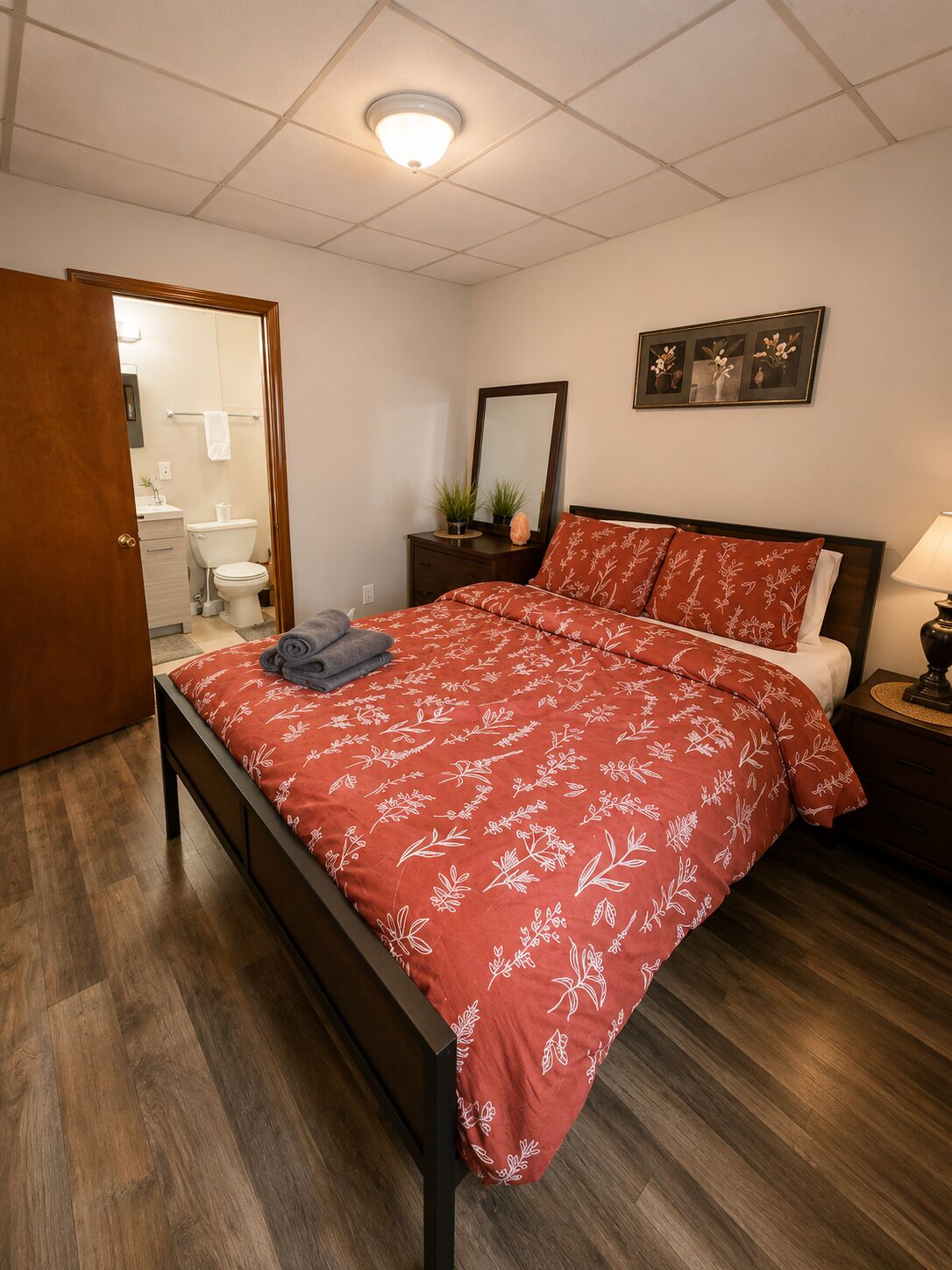 A bedroom with a wooden bed, a bedside table with a lamp, a mirror, and a wall-mounted picture.