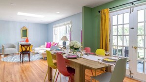 Un comedor con una mesa de madera dispuesta para cuatro personas, sillas coloridas y un sofá al fondo.