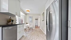 Una cocina moderna con electrodomésticos de acero inoxidable, un comedor con mesa y sillas blancas, y un pasillo que conduce a otra habitación.