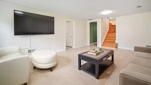 Una sala de estar moderna con un televisor de pantalla plana, un reposapiés redondo blanco, una mesa de centro con libros y una escalera que lleva a una puerta verde.