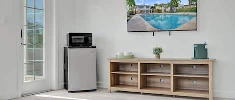 Une pièce avec un grand téléviseur fixé au mur, un petit réfrigérateur, un meuble en bois et une porte avec un panneau de verre.