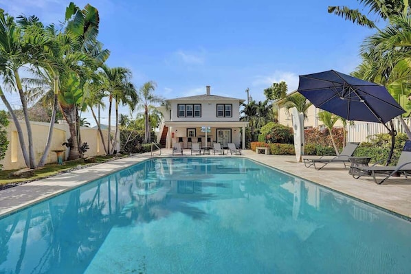 A swimming pool with lounge chairs and an umbrella, surrounded by palm trees and a two-story house.