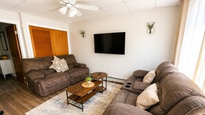 A living room with a brown sofa set, a coffee table, a flat-screen TV, and a ceiling fan. - Cozy 1BR End Cottage W/D • Parking •Walk to Train (Swarthmore)