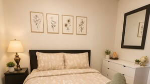 A neatly made bed with floral bedding, a bedside lamp, a nightstand with a plant, and a dresser with a mirror and decorative items. - Cozy 1BR End Cottage W/D • Parking •Walk to Train (Swarthmore)